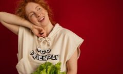 Cheerful woman with red hair in eco-friendly attire holding fresh lettuce against a vibrant red background.